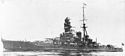Side view of a large dark-gray warship moving at slow speed. Visible are four gun turrets, two funnels, one mast and a prominent forward superstructure.
