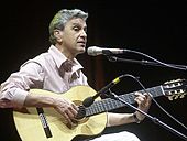 A man sitting behind two microphone stands and playing a guitar