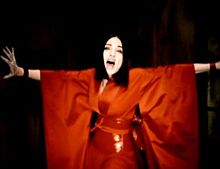 Madonna wearing a red kimono with her hands outstretched to her sides and singing.