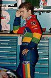 A man his late twenties wearing rainbow colored racing overalls. He has his left hand on his left hip and his right hand leaning against a solid surface.