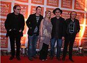 Five people (four men, one woman in the middle) standing next to one another on a red carpet; in the background is a repeated pattern of orange blocks with the text "meteor IRELAND MUSIC AWARDS"