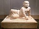 Beige marble statue of a stout young child aged about two years old lying on his left side. The child's head is shaved, his eyes gaze over the viewer's shoulder and his lower body is covered in a draping cloth that hangs limply between his flexed feet. The child supports his torso with his left hand in which he holds an unidentifiable object, he also holds a small bird in his right hand. The sculpture rests on a heavy socle inscribed with barely visible letters spanning the upper part of the socle vertically.