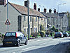 Street scene showing grey stone houses on the left of a road with a few cars.