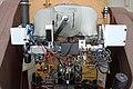 A closeup of electronic devices mounted on the side of the telescope tube, a large brown cylinder. There is a lot of messy cabling, and there are some tubes with thermal insulation. One large piece of hardware, mounted to a circular feature on the side of the telescope tube, is covered with a thermal jacket as well. Written on the jacket with a marker are the names of color channels: "red", "green/photometric", and "blue". On the left and right sides of the picture, the dark brown arms of the equatorial mount fork extend from out of frame at the bottom corners up to the sides of the tube. Below the telescope tube and between the fork arms, the electronic gear shown in the previous picture is visible. Behind it, there is a view to the bottom of the dome, where various items are standing: a work cart, a stepladder, a pedestal fan, etc.