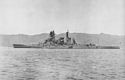 Side view of an anchored dark-gray warship with four gun turrets, two funnels, one mast and a prominent forward superstructure.