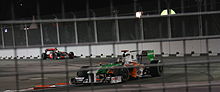 a white, green, and orange F1 car drives down along a closed street at night ahead of a silver F1 car