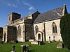 Stone building with square tower to left hand end. Foreground shows gravestones in grass area.