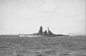 Side view of a dark gray warship at speed. She has two funnels, one mast and a massive forward superstructure.