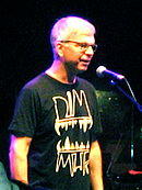 A gray-haired man with glasses and a black shirt standing in front of a microphone