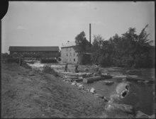 A covered bridge and gristmill. There are also men moving logs on a river.