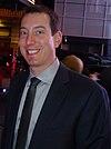 A clean shaven man in his early thirties wearing a black suit, a white shirt and a gray tie