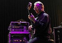 An older man from the side, holding a guitar with an amp to his left under a purple spotlight