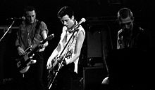 The rock band The Clash performing onstage. Three members are shown. All three have short hair. Two of the members are playing electric guitars.