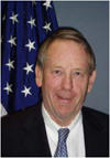 Smiling man with thinning hair wearing a suit and a blue tie with the US flag behind him