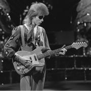 A black and white photo Bowie with longer hair and an eyepatch, holding a guitar and looking down at it