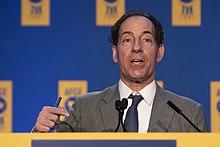 An older man with curly black hair speaking in front of an indoor lectern in front of a blue and yellow backdrop.