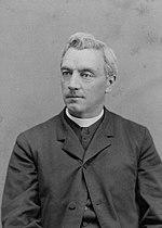 The upper-body of a middle-aged male looking slightly to his right. He wears the black robe and white collar of a Catholic priest