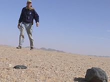 Low angle picture from ground showing sand and a large rock with a man looming a few feet back against a blue sky.