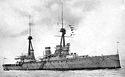 A grey warship with two masts, three funnels, and two gun turrets visible, at anchor