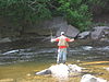 A man in an orange shirt and tan vest fishes in a stream with rapids and large boulders