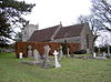 Gray stone building with red tiled roof, partially obscured by a hedge. A square tower is at the far end. The foreground includes several crosses and gravestones.