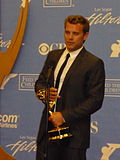 A man with dark hair, wearing a black suit, including a black tie and white T-shirt also holding a gold statuette.