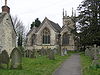 Gray three bay building with arched windows. Tower behind and gravestones in the foreground.