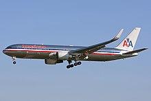 Side quarter view of twin-engine jetliner in flight, with extended gear; bare metal with red, white, and blue cheatlines, as well as two "A"s and a stylized eagle on the tail.