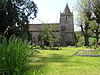 Gray stone building with small square tower and pyramidal roof. Grassy foreground with a cross and gravestones