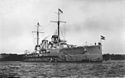 Close front oblique view of a gray warship with two funnels, two visible gun turrets, and two masts
