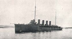 A cruiser sits still in the water with the coastline in the background. Two masts and four funnels sit prominently in the middle of the ship.
