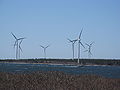 Seven three-blade wind turbines on the shore. A fishing boat is passing by.