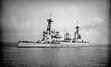 Right rear oblique shot of a large gray warship with two masts, three funnels, and two gun turrets visible. A number of people are visible on her decks.