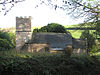Stone building with tiled roof and square tower, surrounded by vegetation.