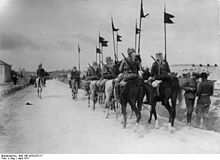 Black-and-white photo of mounted soldiers with middle eastern headwraps, carrying rifles, walking down a road away from the camera