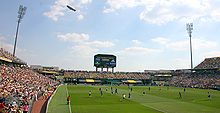 A soccer stadium with a grass field, crowds in the stands, a distant scoreboard, and a blimp flying overhead during the game.