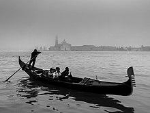 A black-and-white photo of what appears to be a cool, gray day. Four people, indistinct, sit in the middle of a long, thin boat, the gondola, moving to the right. High on the stern (left) stands a man with both hands on a long oar, which disappears into the water to his rear and slightly to his right. In the distant backgound, across the calm water, the buildings of Venice can be made out, standing in the mist.