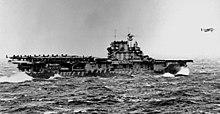 Black and white photo of a flat-decked ship at sea. It is carrying aircraft on the rear of its deck, and one plane is flying immediately in front of the ship.