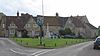 Street scene. Triangular area of grass with village sign on wooden post and stone cross behind. Stone houses with tiled roofs n the background.