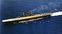 left front oblique aerial view of a dark-gray aircraft carrier at sea. Her flight deck is beige colored and has a dozen or so aircraft at the stern.