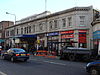 A pale two-storey stone building, with gold coloured letters at the top reading "METROPOLITAN RAILWAY" and "PADDINGTON STATION", windows on the first floor. On the ground floor there are shops either side of an entrance with a canopy with rectanglar blue signs reading "PADDINGTON STATION". There are cars and a small truck in the road and people are walking on the pavement.