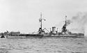 Side view of a gray warship with two masts and two visible gun turrets at anchor. Smoke is rising from one of the two funnels.
