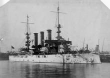 A white battleship with three smokestacks and two tall masts sitting in port.