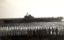 Black and white photo of men standing in formation on the deck of a ship looking at another ship which is sailing nearby